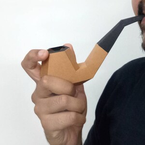 Peut inclure: Une personne tient une pipe en carton marron avec un embout noir. La pipe est de forme traditionnelle.