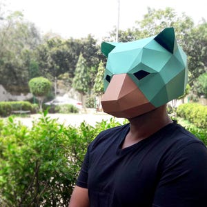 May include: A person wearing a green and brown paper mache animal mask. The mask has a geometric design and resembles a bear.