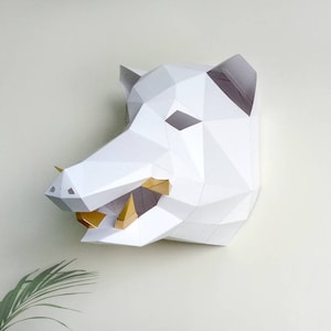 May include: White papercraft sculpture of a wild boar head with gold paper tusks. The sculpture is mounted on a white wall.