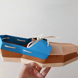 Op de afbeelding: Een papieren model van een bruine en blauwe bootschoen met witte veters. De schoen is gemaakt van gevouwen papier en heeft een realistisch uiterlijk.