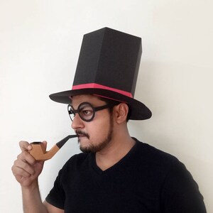 Può includere: Una persona che indossa un cappello a cilindro nero con una fascia rosa e occhiali rotondi, tiene una pipa marrone.