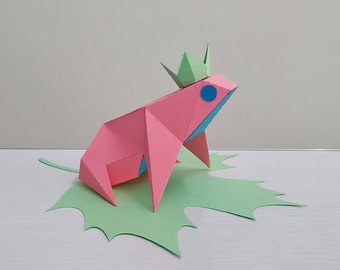 May include: A pink origami frog wearing a green paper crown sits on a green paper leaf. The frog has blue eyes.