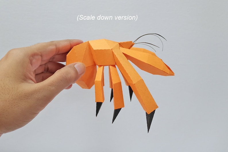May include: An orange papercraft model of a crab with black claws and antennae. The crab is being held in someone's hand.