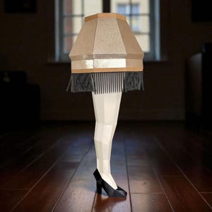 May include: A quirky lamp in the shape of a leg, featuring a beige lampshade, black fringe, and a high-heeled shoe. The leg is white, and the shoe is black. The lamp is on a dark wood floor.