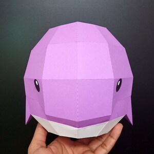 May include: A purple papercraft whale head with white accents. The whale has black eyes and a simple, geometric design.
