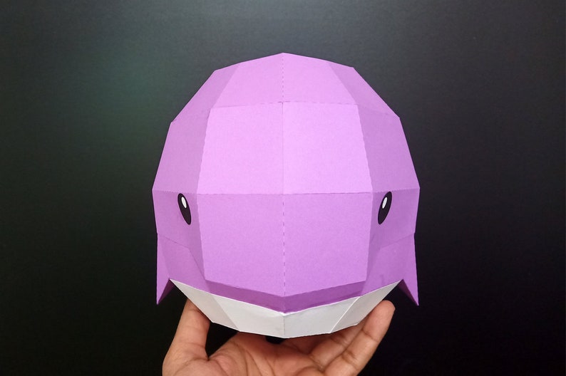 May include: A purple papercraft whale head with white accents. The whale has black eyes and a simple, geometric design.