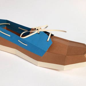 Op de afbeelding: Een papieren model van een bruine en blauwe bootschoen met witte veters. De schoen is gemaakt van gevouwen papier en heeft een realistische uitstraling.