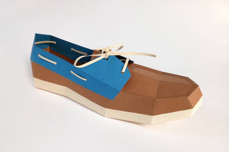 Op de afbeelding: Een papieren model van een bruine en blauwe bootschoen met witte veters. De schoen is gemaakt van gevouwen papier en heeft een realistische uitstraling.