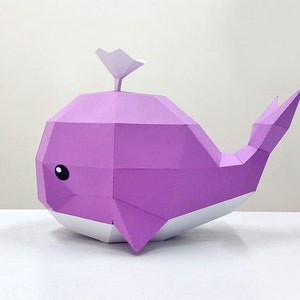 May include: A purple papercraft whale with a white fin and belly. The whale has a simple, cartoon-like face with black eyes.