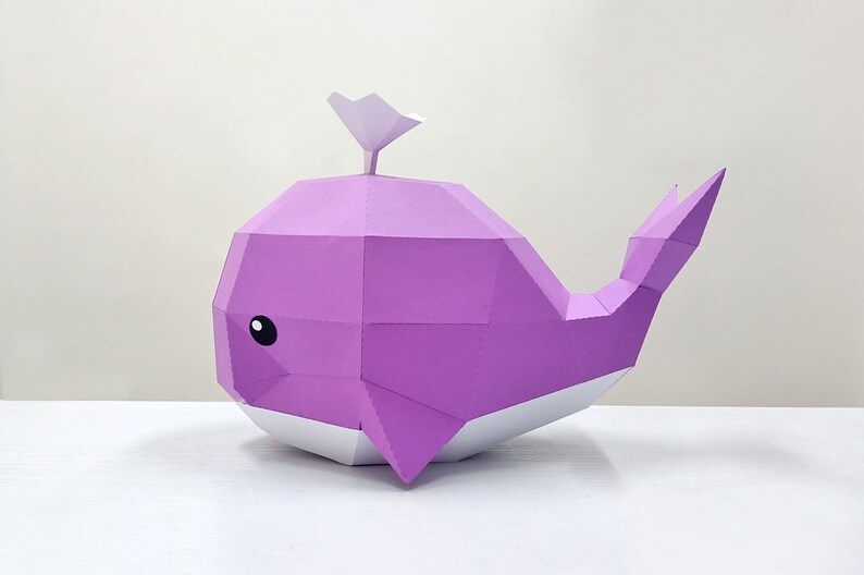 May include: A purple papercraft whale with a white fin and belly. The whale has a simple, cartoon-like face with black eyes.