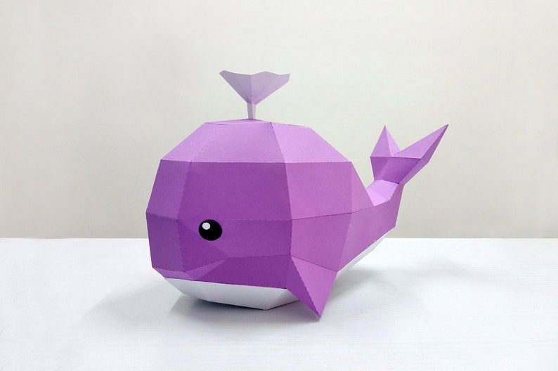 May include: A purple papercraft whale with a white belly and a small purple fin on top of its head. The whale has a simple, cartoon-like face with a black eye.