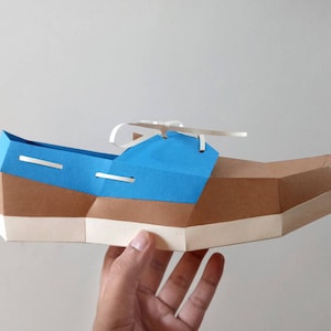 Op de afbeelding: Een papieren model van een blauwe en bruine bootschoen met witte veters. De schoen wordt in een hand gehouden.