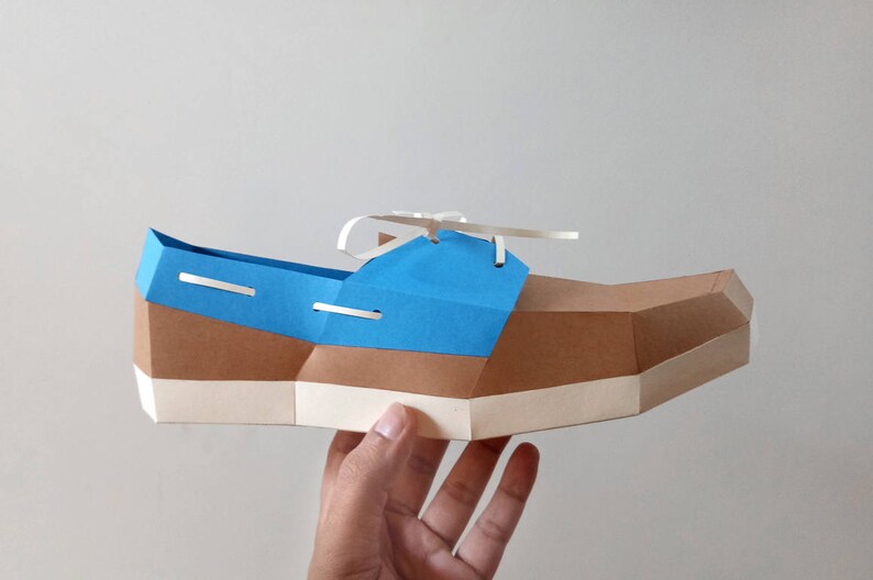 Op de afbeelding: Een papieren model van een blauwe en bruine bootschoen met witte veters. De schoen wordt in een hand gehouden.