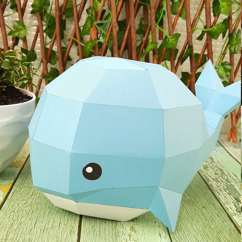 May include: A light blue papercraft whale with a white belly and black eyes. The whale is made of folded paper and has a 3D effect.