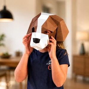 May include: A low-poly paper craft dog mask featuring a geometric design with brown, white, and black panels. The mask covers the head and face, showcasing a stylized canine appearance with a white muzzle, a black nose, and brown ears.