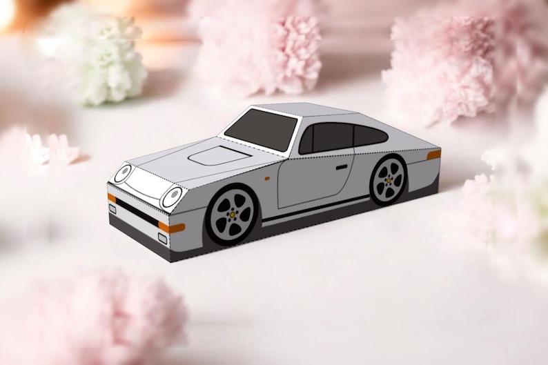 May include: A gray paper model of a sports car with black wheels and a black windshield. The car has a sleek, angular design with a low profile. The background features soft pink and white flowers.