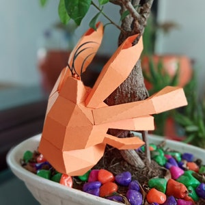 May include: An orange papercraft crab sculpture perched on a small potted tree with colourful stones. The crab has a detailed, geometric design and is made of folded paper.