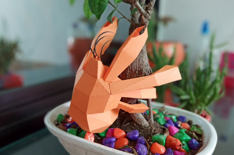 May include: An orange papercraft crab sculpture perched on a small potted tree with colourful stones. The crab has a detailed, geometric design and is made of folded paper.