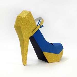 May include: A gold and blue papercraft high heel shoe. The shoe has a gold glitter finish and a blue strap.