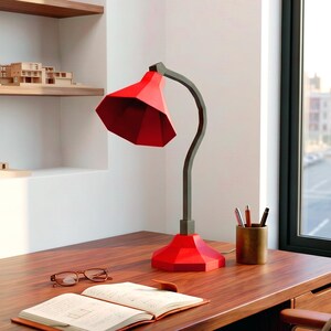May include: A red geometric desk lamp with a dark gray stem and base sits on a wooden desk. A notebook, glasses, and pen holder are also on the desk. Architectural models are on shelves in the background.
