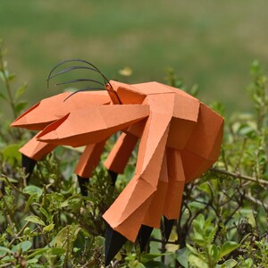 May include: An orange papercraft hermit crab with black antennae and legs. The crab is posed on a green bush.