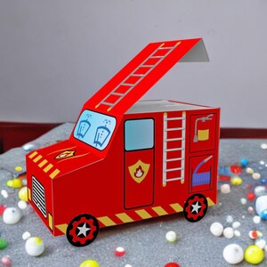 Könnte beinhalten: Ein roter Karton in Form eines Feuerwehrautos mit einem Klappdeckel. Die Schachtel hat eine Leiter an der Seite und einen Feuerwehrschlauch auf der Rückseite. Die Schachtel ist mit einem Feuerwehrauto-Design verziert.