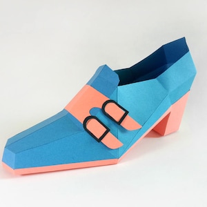 May include: A blue and pink papercraft shoe with two black buckles. The shoe has a high heel and a pointed toe.