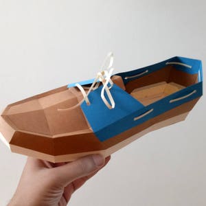 Op de afbeelding: Een papieren model van een bruine en blauwe schoen met witte veters. De schoen is gemaakt van karton en heeft een geometrisch ontwerp.