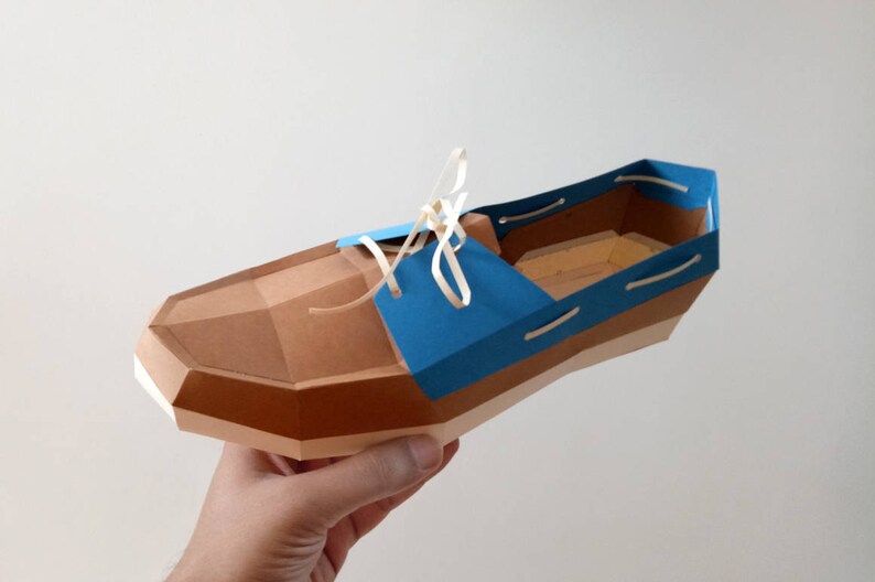 Op de afbeelding: Een papieren model van een bruine en blauwe schoen met witte veters. De schoen is gemaakt van karton en heeft een geometrisch ontwerp.