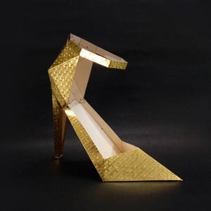 May include: A gold-colored, geometrically shaped high heel shoe. The shoe is made of paper and has a textured surface.