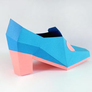 May include: A blue and pink origami shoe with a high heel. The shoe is made of paper and has a geometric design.