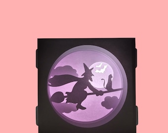May include: A black Halloween-themed paper cut-out scene. A witch on a broom flies in front of a full moon with bats. A black cat sits on a branch. The scene is set inside a black square frame with a pink background.