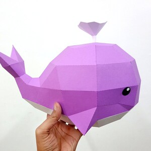 May include: A purple papercraft whale with a white underside and black eyes. The whale has a small fin on its back.