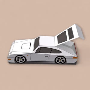 May include: A white and gray paper model of a sports car with a raised rear engine cover. The car has black wheels and windows, and is set against a neutral background. The model is a detailed representation of a classic car.
