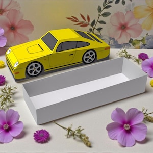 Puede incluir: Un modelo de papel amarillo de coche deportivo con ruedas negras y una caja rectangular blanca. El coche está colocado encima de la caja. La escena está decorada con flores rosas, moradas y amarillas y vegetación.