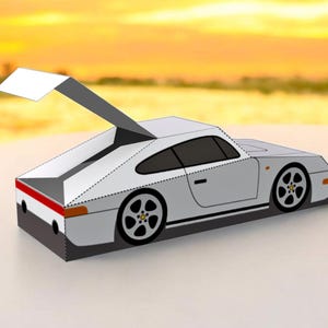 May include: A white paper model of a sports car with black wheels and a raised rear spoiler. The car has black windows and a red stripe on the back. The background is a blurred sunset.