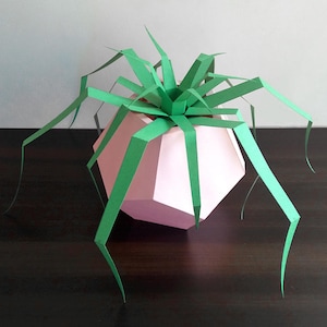 May include: A pink paper geometric planter with a green paper plant. The plant has long, thin leaves that extend out from the planter.