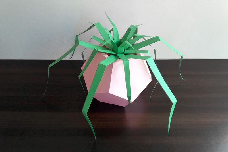 May include: A pink paper geometric planter with a green paper plant. The plant has long, thin leaves that extend out from the planter.