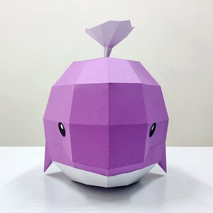 May include: A purple papercraft whale with a white belly and black eyes. The whale has a small, pointed fin on top of its head.