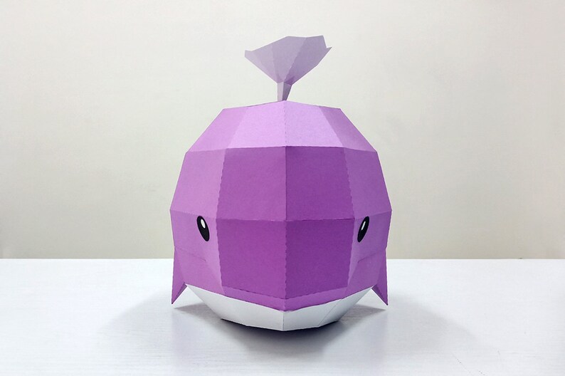 May include: A purple papercraft whale with a white belly and black eyes. The whale has a small, pointed fin on top of its head.
