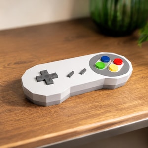 May include: A white and gray retro-style game controller with a cross-shaped directional pad and colored buttons. The controller is on a wooden surface, with books and greenery in the background. A nostalgic item for gamers.