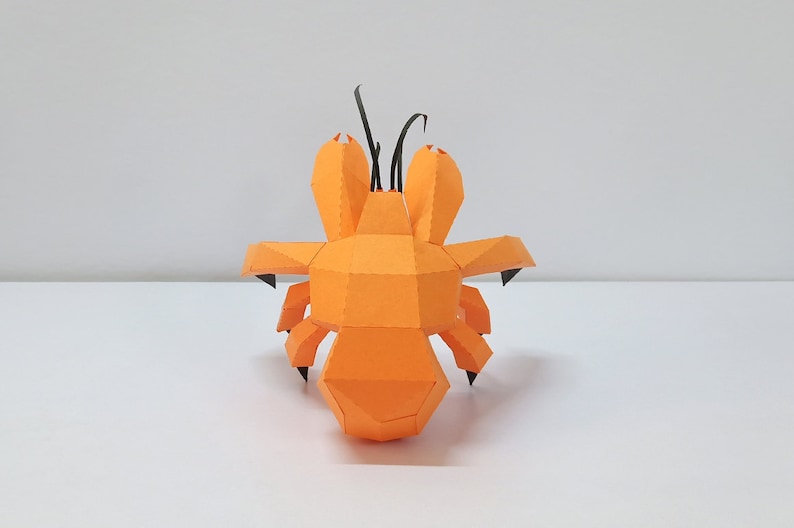 May include: An orange papercraft model of a hermit crab with black antennae. The model is made of folded paper and has a realistic appearance.