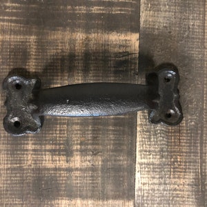 May include: Black cast iron door handle with a decorative design. The handle is about 6 inches long and has a smooth, slightly textured surface.