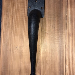 May include: Black cast iron door handle with a curved design and a square back plate. The handle is mounted on a wooden surface and comes with screws in a plastic bag.