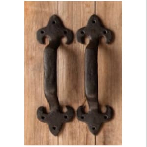 May include: Two black metal door pulls with a fleur-de-lis design. The pulls are mounted on a wooden surface.