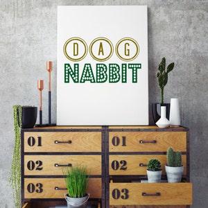 May include: A white poster with the text "DAG NABBIT" in green letters with gold circles around the letters D, A, and G. The poster is hanging on a gray concrete wall above a wooden dresser with five drawers. The dresser has black metal accents and each drawer has a number on it.