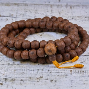 May include: A brown wooden mala bead necklace with a tassel made of yellow cord. The beads are round and have a textured surface. The necklace is lying on a white wooden surface.