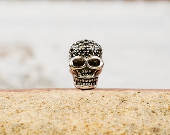 Puede incluir: Un amuleto de calavera de plata con detalles de cristal negro. La calavera tiene un diseño detallado con cuencas de los ojos y dientes.