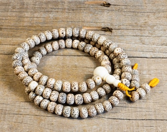 May include: A mala bead necklace with speckled, off-white beads and a large, smooth, white bead. The necklace is strung on a yellow cord and rests on a wooden surface. The beads have dark brown spots.
