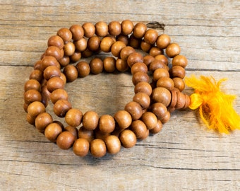 May include: A brown wooden mala bead necklace with a tassel made of orange fabric. The necklace is made of round beads and is lying on a wooden surface.